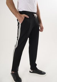 Svarta joggerbyxor med vita sidostreck, dragsko i midjan och fickor, tillsammans med svarta sneakers. Slät textur och figurnära design.