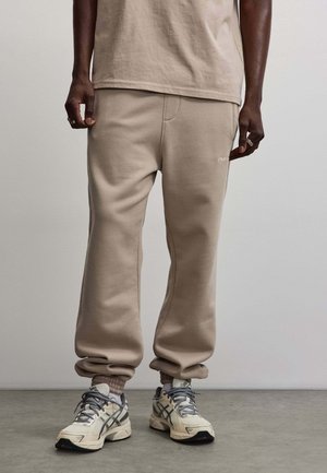 Parte inferiore del corpo di una persona che indossa pantaloni della tuta beige, maglietta beige coordinata, calzini grigi e scarpe da corsa bianche, in piedi su un pavimento grigio.