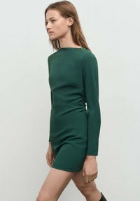 Vestido ajustado de manga larga en tejido verde oscuro con fruncidos laterales, escote redondo y un dobladillo corto. Textura suave y silueta elegante.