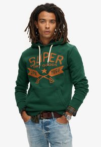 Sudadera verde con un gran logo naranja de "SUPER DRY GOODS", con un bolsillo frontal y capucha con cordón. Lleva puesta sobre una camisa de cuadros y unos jeans desgastados.