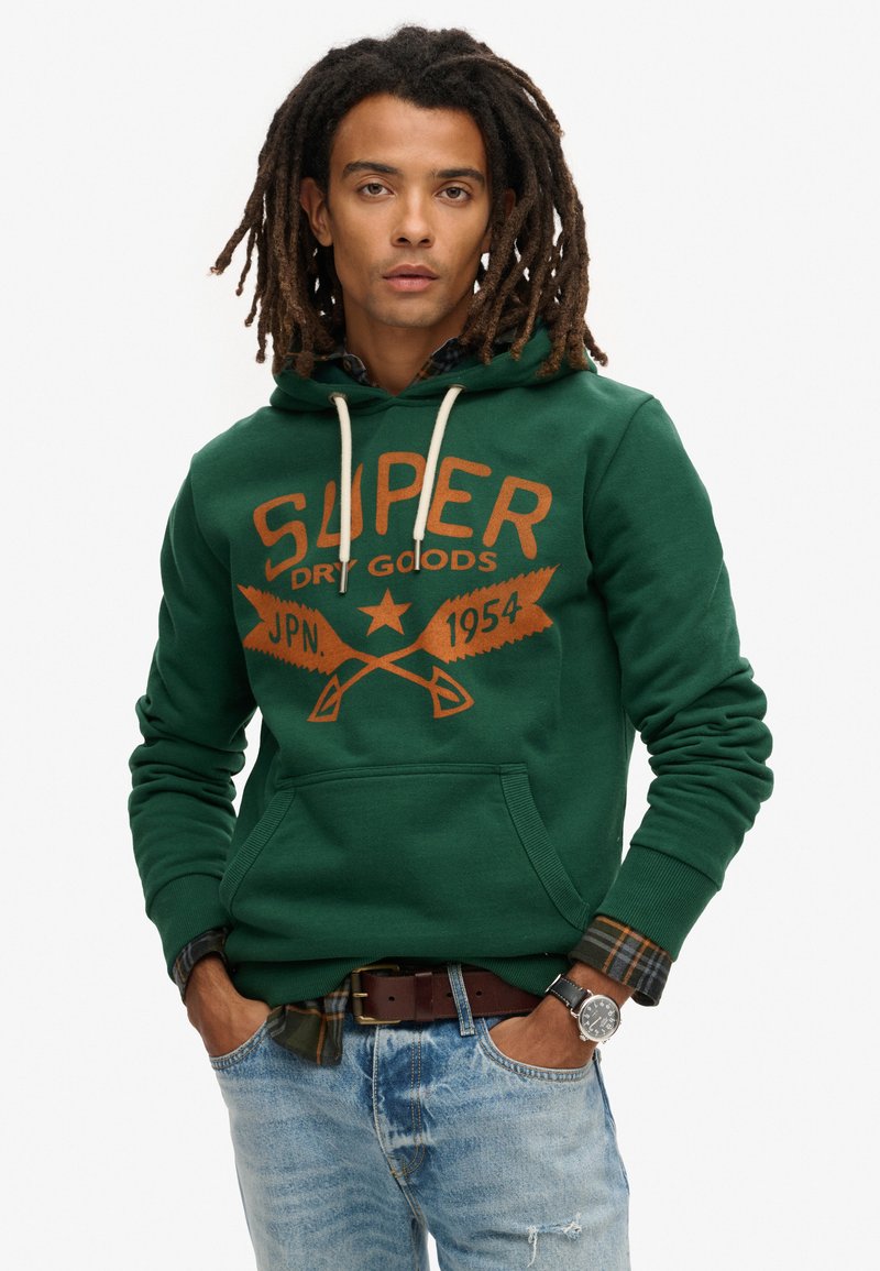 Sudadera verde con un gran logo naranja de "SUPER DRY GOODS", con un bolsillo frontal y capucha con cordón. Lleva puesta sobre una camisa de cuadros y unos jeans desgastados.