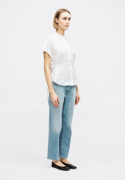 Γυναίκα που στέκεται πλαγίως φορώντας λευκό πουκάμισο με κοντό μανίκι και κουμπιά, τζιν παντελόνι ίσιας γραμμής ανοιχτού μπλε χρώματος και μαύρα επίπεδα παπούτσια σε λευκό φόντο.