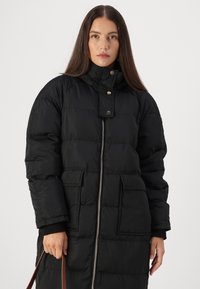 Lang sorte pufferjakke med høj krave, trykknaplukning, frontlynlås og to store lommer. Glat tekstur med et syet mønster.