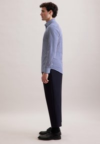Uomo in profilo che indossa una camicia a righe blu e bianche con bottoni, pantaloni scuri, calze nere e scarpe eleganti nere.