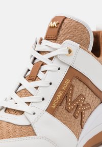 Weißer Sneaker mit geflochtenem, braunem Material, goldenen Akzenten, dekorativem Logo und weißen Schnürsenkeln. Glatte Textur mit kontrastierenden Einsätzen.