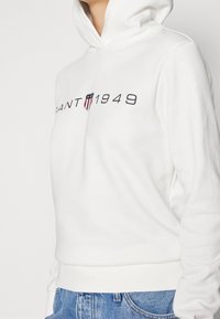 Sudadera blanca con capucha de material suave que presenta una impresión gráfica con "CANT 1949" y un diseño de escudo. Puños y cinturilla ajustados.