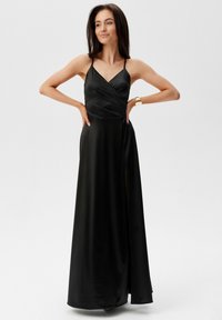 Černé saténové maxi šaty s obepnutým designem, tenkými ramínky a bočním rozparkem. Hladká textura s plynulou siluetou a jemným leskem.