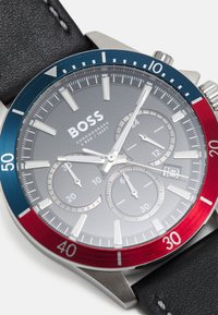 Reloj cronógrafo plateado con bisel azul y rojo, esfera gris, tres subesferas y correa de cuero negra. Presenta índices y manecillas plateadas.