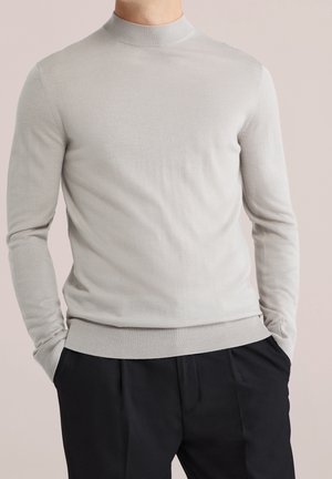 Man wearing light gray long-sleeve turtleneck sweater and black pants with hands in pockets against neutral background.