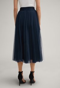 Jupe en tulle bleu marine avec une taille élastique noire, longueur cheville, design superposé et texture transparente. Associée à des chaussures à talons noirs.