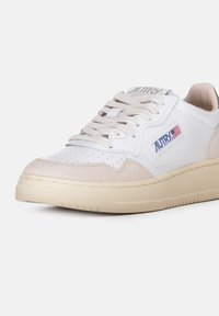 Sneaker bianca con accenti in suede beige, punta perforata, lacci piatti e una patch con logo. Suola in gomma testurizzata con scanalature per una trazione distintiva.