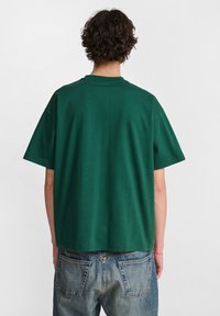 Zielona bawełniana koszulka z krótkim rękawem, okrągłym dekoltem i luźnym krojem. Widok z tyłu pokazujący gładką fakturę i minimalistyczne detale.