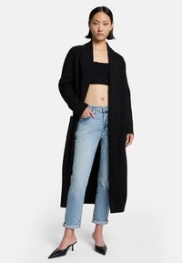 Cappotto di lana nero con maniche lunghe, davanti aperto e tasche, abbinato a jeans azzurri con risvolto e mules nere con tacco.