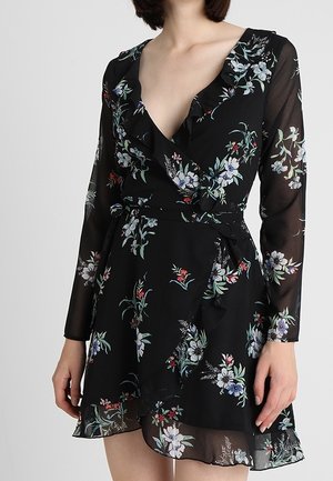 Femme portant une robe portefeuille noire à motifs floraux, manches longues en voile transparent et ourlet à volants, debout devant un fond uni.