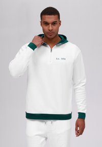 Hvit zip-up hettejakke med grønn hette og mansjetter. Har teksten "Est. 1958" i grønn brodering. Laget av et glatt, teksturert stoff.