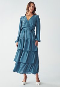 Gaun teal panjang dengan desain lilit, lengan panjang, ruffles bertingkat, dan pinggang yang terdefinisi. Dipadukan dengan sepatu hak tinggi putih.