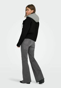 Žena u crnoj jakni sivi duksericom ispod i sivim trapericama sa širokim nogavicama, stoji okrenuta leđima.