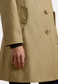 Cappotto beige doppiopetto con due bottoni, tasche a taglio e una texture liscia. Maniche con risvolto e dettagli di cucitura sottili.