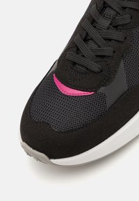 Musta lenkkari, jossa on verkkokangaskappaleita päällisessä, mokkasiinisomisteita ja vaaleanpunaisia yksityiskohtia. Siinä on kuvioidut kengännauhat ja valkoinen kumipohja.