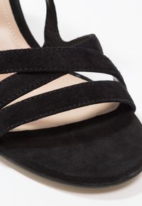Gros plan d'une sandale en daim noir avec deux brides horizontales et une semelle intérieure beige sur fond blanc.