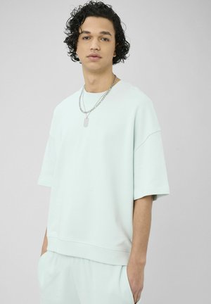 Jeune homme aux cheveux bouclés portant une chemise oversize vert menthe claire et un short assorti, accessoirisés avec des chaînes argentées superposées.