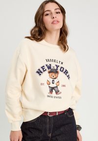 Sweat-shirt crème avec un graphique d'ours portant une casquette et une chemise, avec les inscriptions "BROOKLYN" et "NEW-YORK". Manches et bas côtelés.