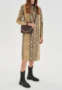 Mujer con un abrigo de estampado de serpiente hasta la rodilla, botas negras hasta media pantorrilla y llevando un bolso cruzado marrón oscuro con detalles de cadena dorada.