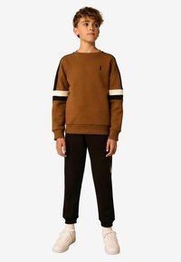 Sweat-shirt marron avec des rayures noires et blanches sur les manches, pantalon noir et baskets blanches ; présente un petit logo sur la poitrine.