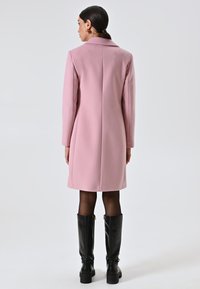 Donna con capelli scuri raccolti in un basso chignon, che indossa un cappotto rosa al ginocchio, collant neri e stivali neri fino al ginocchio, in piedi contro uno sfondo chiaro e semplice.