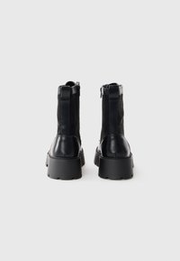Une paire de bottes plateformes en cuir noir et en daim avec fermeture éclair à l'arrière et semelle épaisse, présentée de l'arrière sur fond blanc.