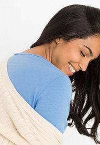 Camicetta azzurro chiaro a maniche corte indossata sotto un cardigan lavorato a maglia color crema, con una texture a coste e un fit casual. I capelli sono scuri e mossi.
