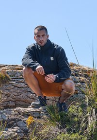 Veste polaire noire avec poche zippée, shorts orange et baskets noires ; l'arrière-plan présente un terrain rocheux et de la végétation.