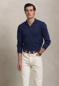 Camisa polo de manga longa azul-marinho com colarinho, usada com jeans brancos e um cinto castanho; modelo segurando um relógio de pulso e de pé contra um fundo suave.