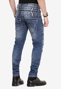 Herren Jeans aus blauem Denim mit schmaler Passform, Mehrfachtaschen-Design, verwaschenen Akzenten und auffälligen Riemendetails am Bund.