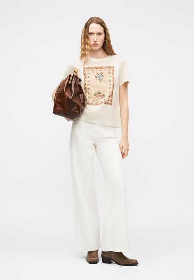Γυναίκα με κυματιστά μαλλιά που φοράει κρέμ τοπ με φλοράλ σχέδιο, λευκό παντελόνι φαρδύ στο πόδι, καφέ μπότες, κρατώντας μια μεγάλη καφέ δερμάτινη τσάντα.