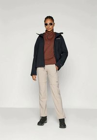 Giacca nera impermeabile, maglione in pile color ruggine, pantaloni beige con zip e scarponi da trekking neri. Gli accessori includono occhiali da sole.