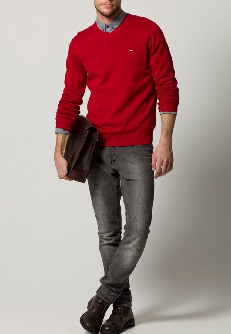 Homme portant un pull rouge par-dessus une chemise grise, un jean gris délavé et des bottes marron foncé, tenant une sacoche en cuir marron dans la main gauche.