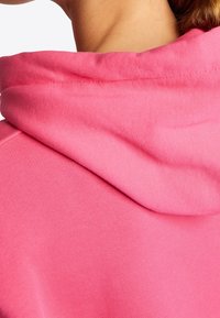 Różowa bluza z kapturem o miękkiej teksturze; posiada złożony kaptur oraz widoczne szwy wzdłuż dekoltu i szwu ramion.