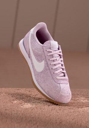 Jasnofioletowy zamszowy sneaker Nike z białym logo Swoosh, sznurowadłami i gumową podeszwą na dopasowanej jasnobrązowej teksturowanej powierzchni.