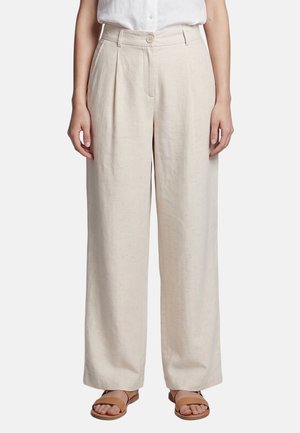 Frau trägt beige Leinenhose mit hoher Taille und weitem Bein mit Vorderfalten, kombiniert mit einem weißen, eingesteckten Hemd und braunen Sandalen.