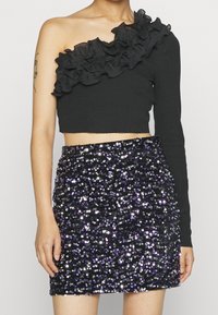 Mustasosalla musta ribbed crop-paita yhdellä hihalla ja röyhelöyksityiskohdilla, yhdistetty tiukkaan kimaltavaan purppuraan paljetein koristeltuun mini-hameeseen.