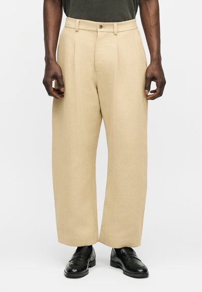 Pantalon beige à jambes larges avec bouton et passants pour ceinture, porté avec des chaussures en cuir noir et une chemise foncée rentrée.