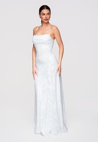 Femme portant une robe blanche sans manches, longue jusqu'au sol, avec une texture florale subtile, debout, mains détendues, regardant sur le côté.