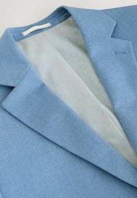 Světle modré texturované sako s výstřihem se zářizy a jemně vzorovanou světle šedou vnitřní podšívkou, zobrazené na bílém pozadí.