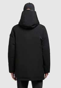Hombre con un abrigo de invierno negro con capucha y una gorra negra de béisbol, visto desde atrás frente a un fondo sencillo.