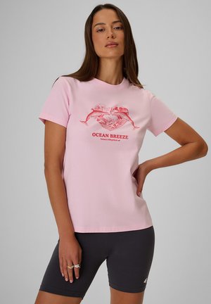 Kobieta w różowym t-shircie z czerwonym delfinem i sercem oraz napisem ""Ocean Breeze"", w połączeniu z czarnymi spodenkami rowerowymi.