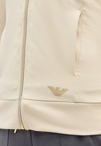 Detailný záber na béžovú bundu na zips s bočným vreckom a malými zlatými nitmi pri leme nad tmavými nohavicami.