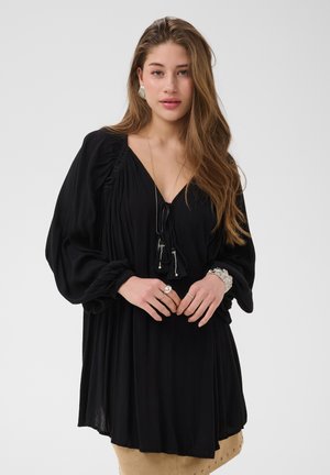Jeune femme aux longs cheveux bruns porte une robe noire ample avec des manches froncées, des bijoux argentés et une jupe beige ornée de clous métalliques.