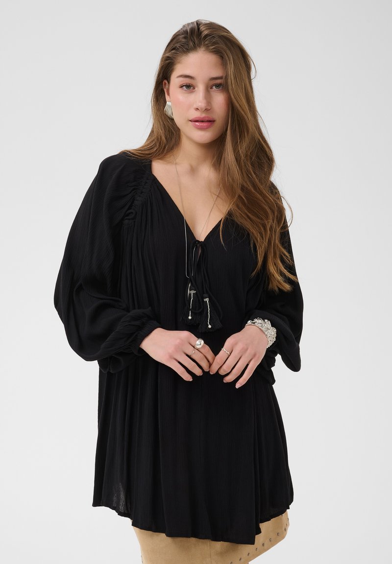 Jeune femme aux longs cheveux bruns porte une robe noire ample avec des manches froncées, des bijoux argentés et une jupe beige ornée de clous métalliques.