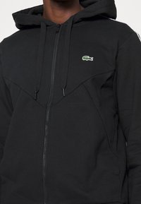 Svart dragkedja hoodie gjord av bomullsblandning, med en känguruficka, texturerad tyg och grön broderad krokodil-logga på bröstet.